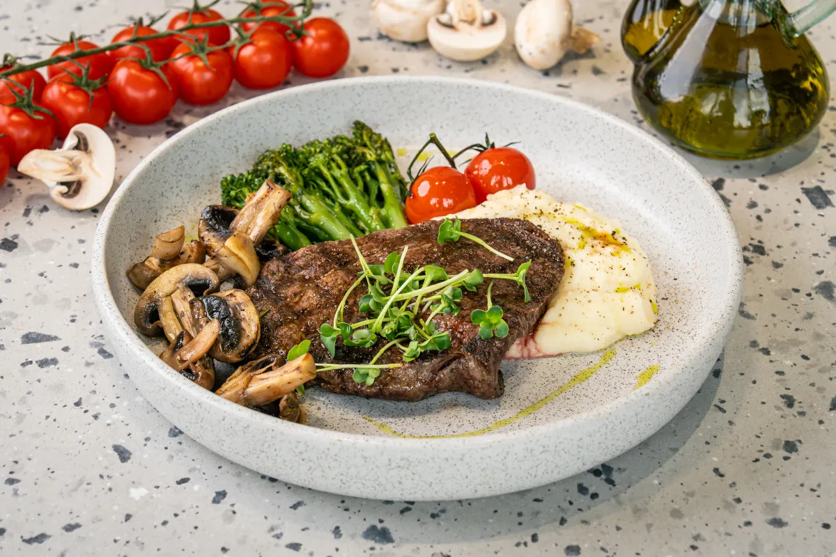 Seared beef fillet with sautéed mushrooms, broccolini, vine tomatoes and a smear of mash, finished with cress, in a speckled bowl