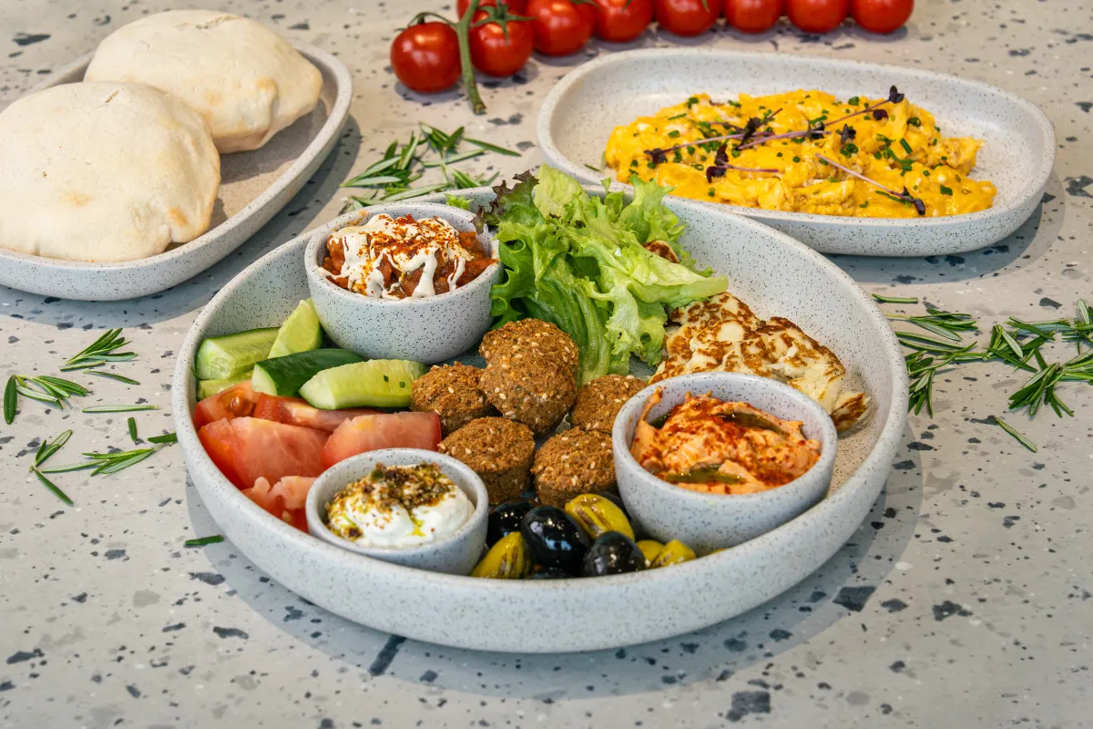 Speckled tiered platter of falafel, olives, tomato and cucumber around pots of foul, labneh and hummus, with scrambled eggs and pita alongside