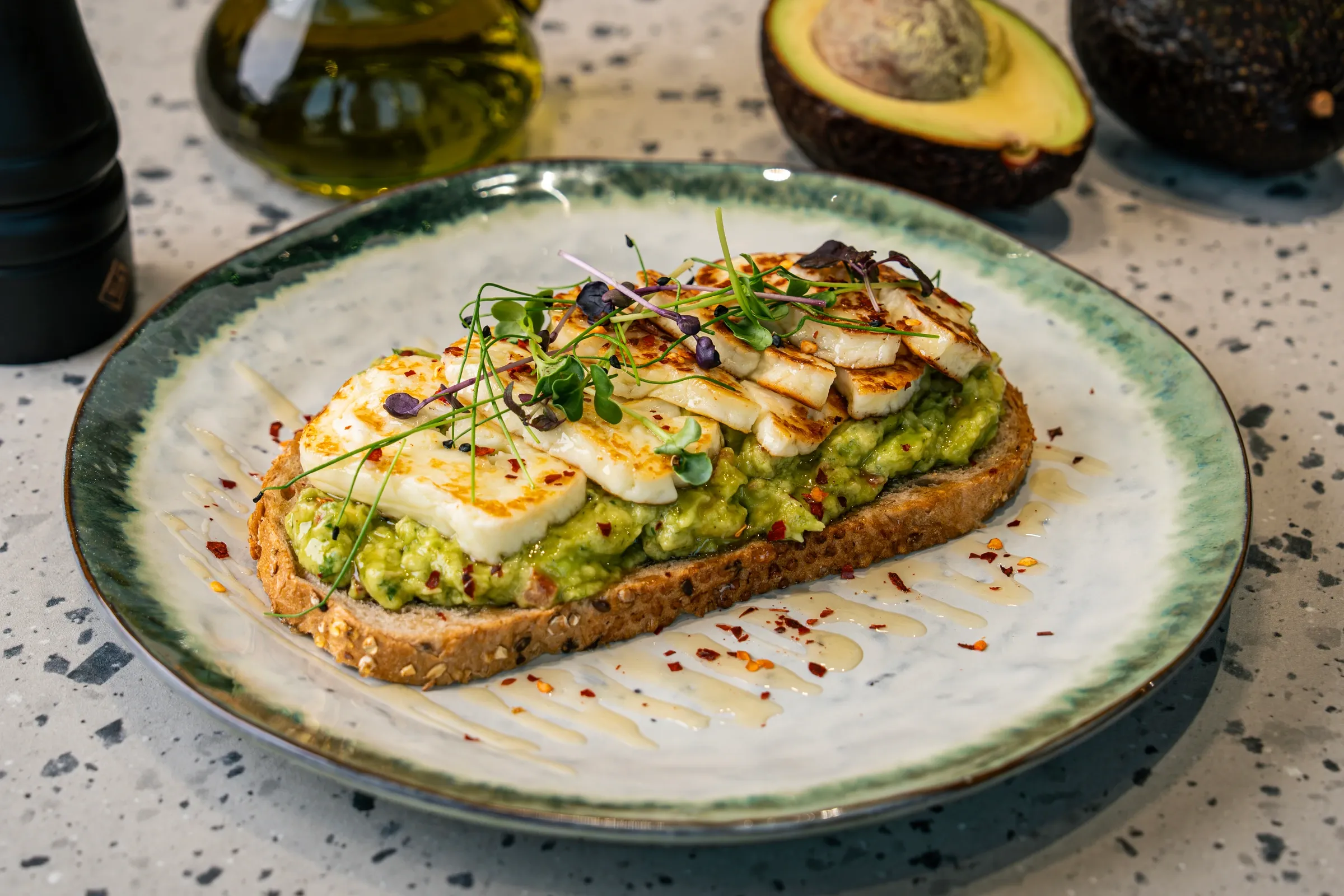Griddled halloumi slices fanned over smashed avocado on seeded sourdough, with microgreens, chilli flakes and a honey zigzag on the plate