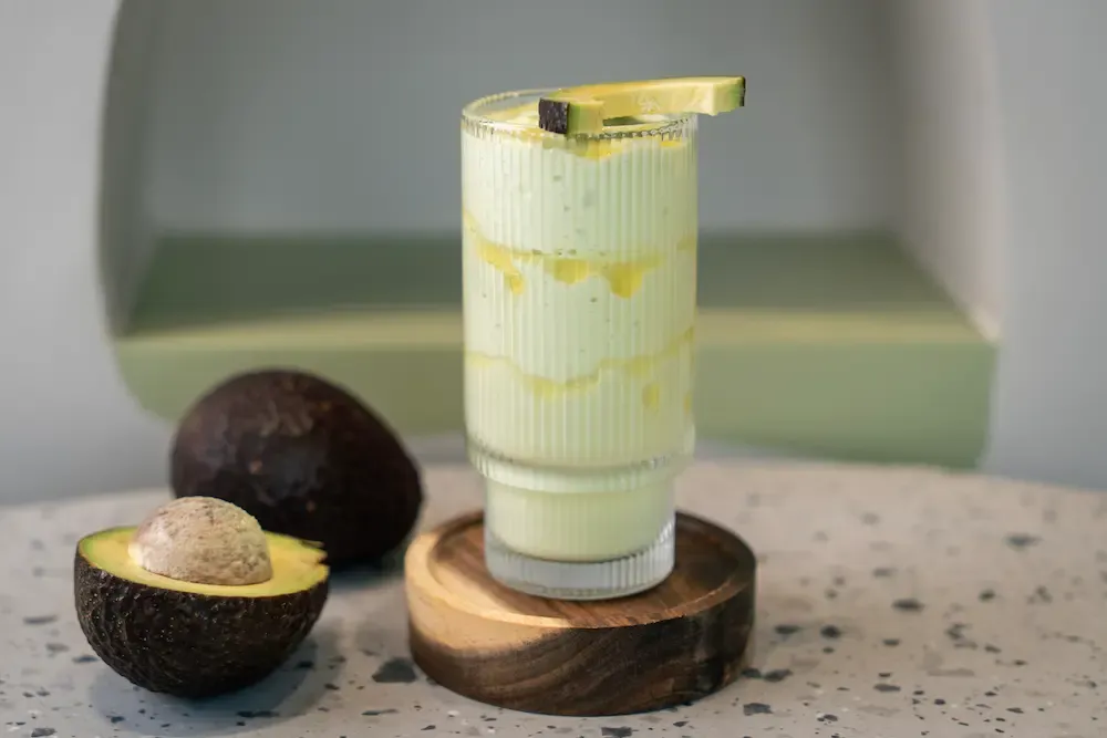 Pale green smoothie in a ribbed glass with honey streaking down the inside, an avocado wedge on the rim and a halved avocado beside it