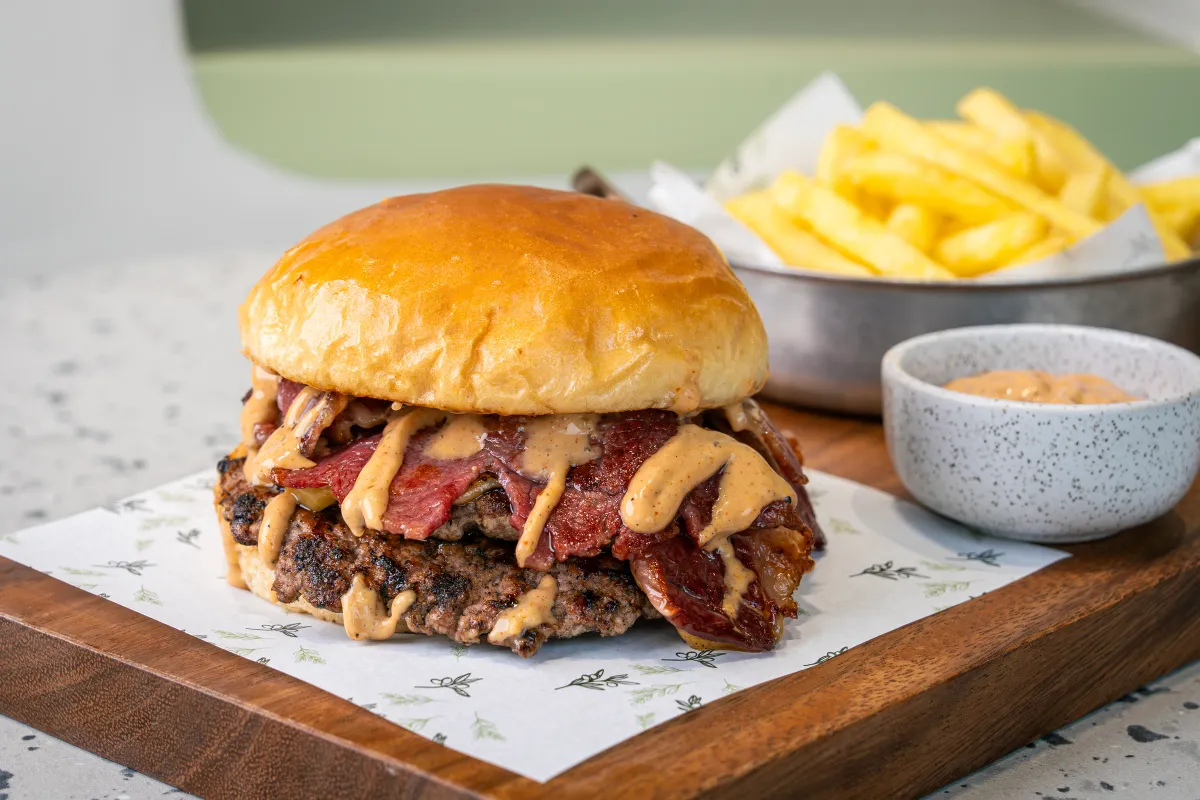 Thick charred patty piled with folded strips of red beef bacon and zigzags of tan sauce under a glossy brioche lid, chips alongside