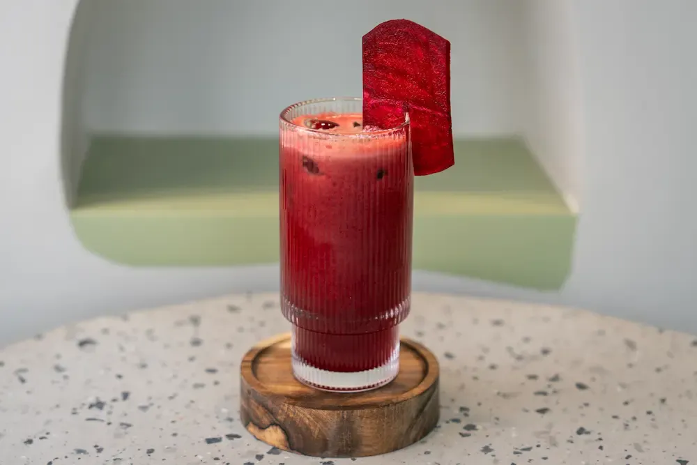 Deep crimson juice in a tall ribbed glass on a round wooden coaster, a thin dried beetroot slice clipped to the rim