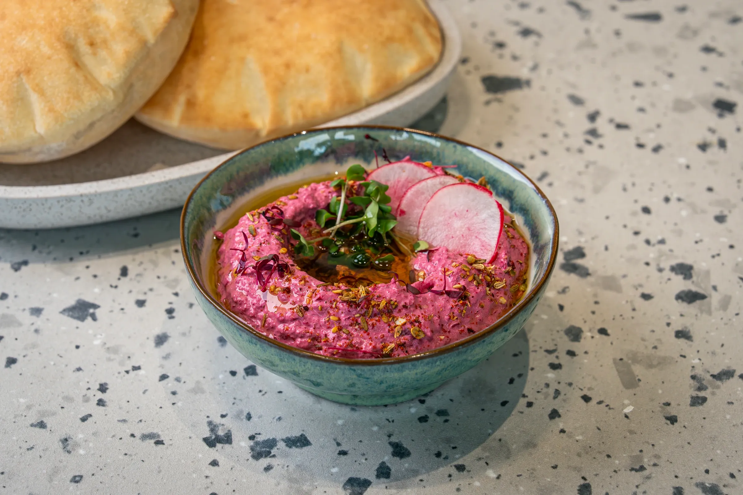 Bright pink beetroot dip ringed with olive oil in a green glazed bowl, topped with radish slices, micro herbs and spice, pitta behind