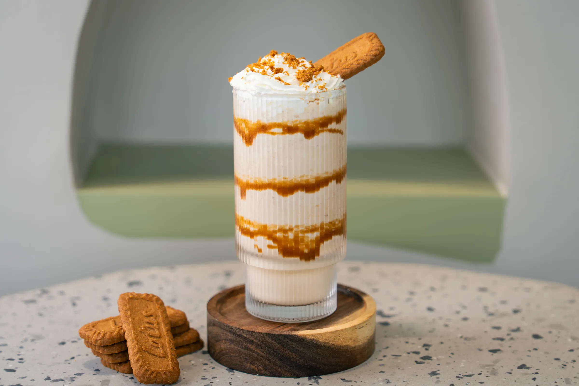 Ribbed glass of cream shake ringed with caramel sauce, whipped cream and biscuit crumb on top, a Lotus biscuit standing in it