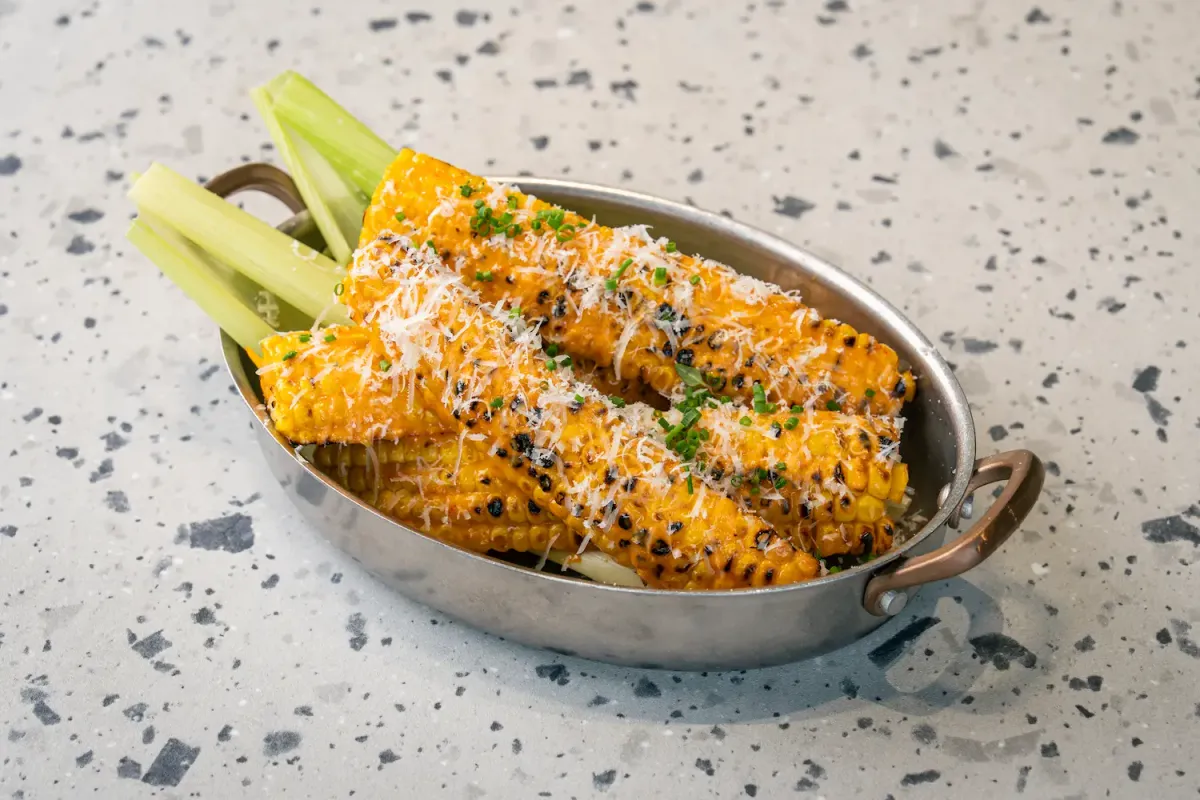 Charred corn ribs under a heavy shower of grated Parmesan and chopped chives, packed into a small steel pan with copper handles