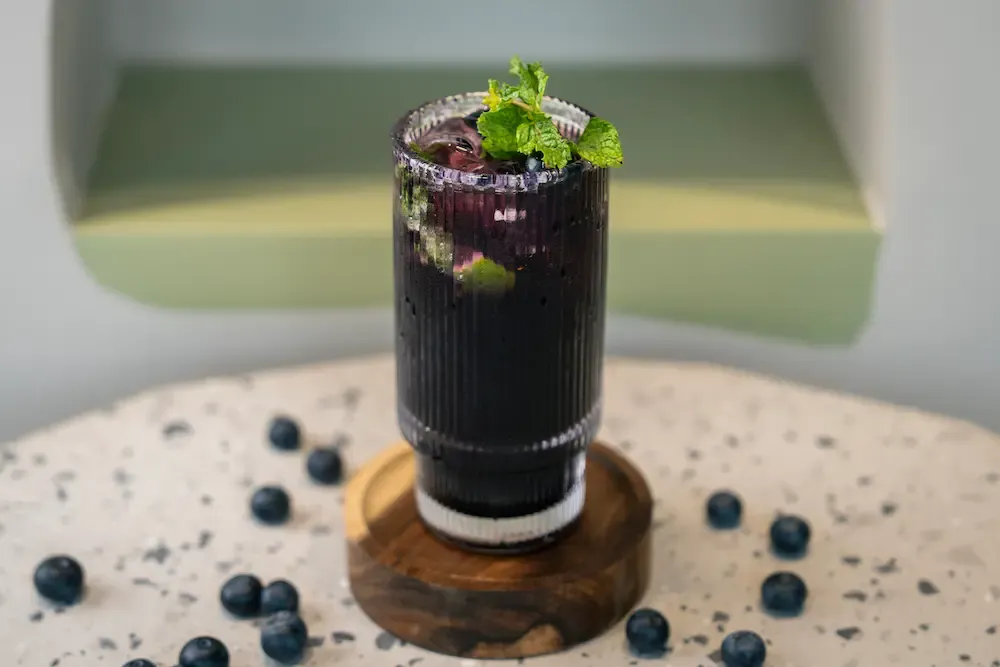 Deep purple drink in a tall ribbed glass on a wooden coaster, mint and blueberries on top, loose berries scattered around the base