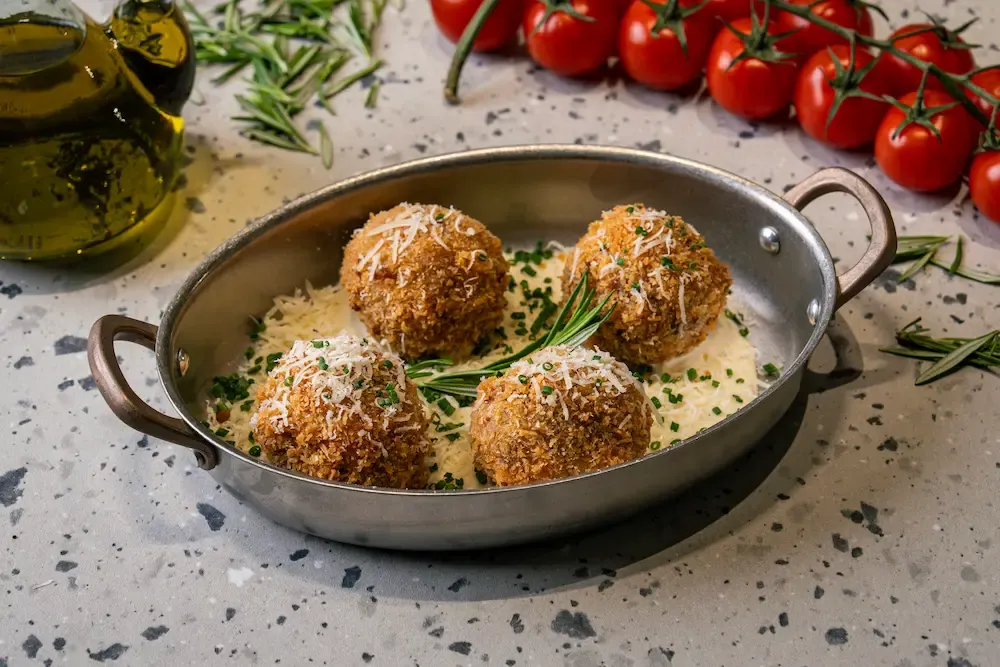 Four panko-crusted rice balls sitting in a pool of cream sauce in a steel pan, scattered with shaved cheese, chives and rosemary