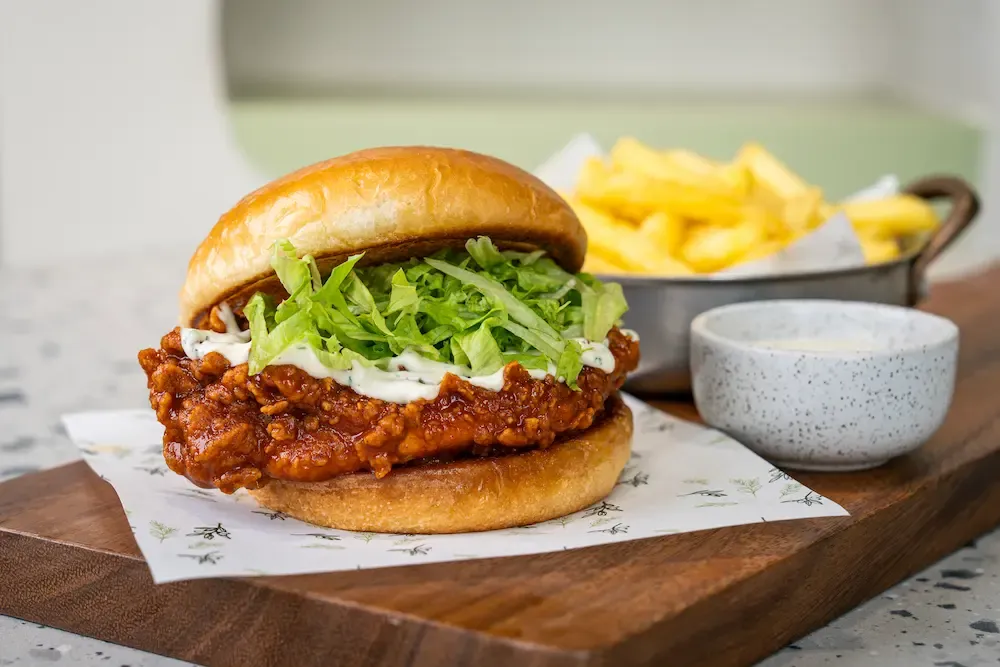 Fried chicken fillet in a deep red glaze under shredded lettuce and a white ranch drizzle, brioche bun, chips and a dip pot behind