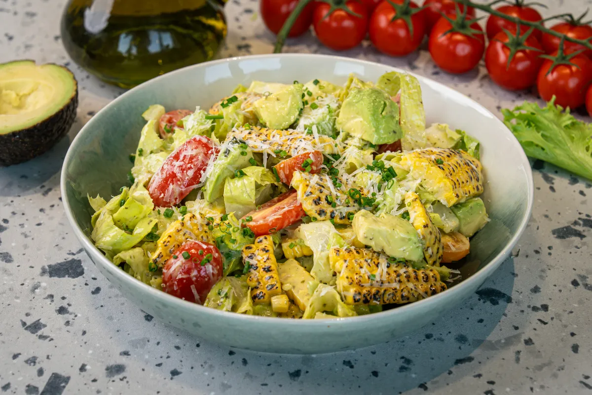 Chopped lettuce, avocado and halved cherry tomatoes with charred corn ribs and grated cheese heaped in a pale green bowl
