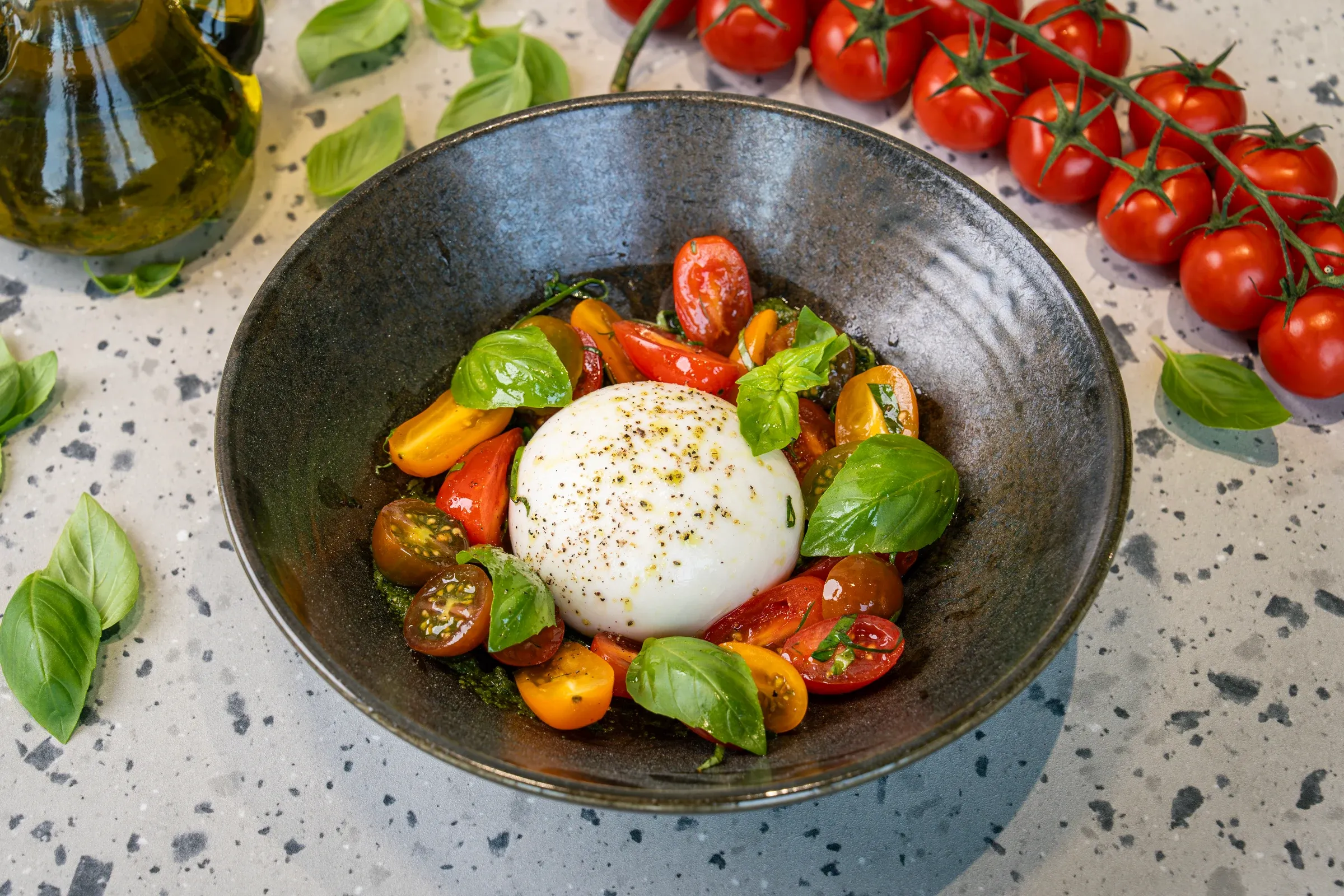 Whole burrata dusted with cracked pepper in a dark bowl, ringed with red and yellow tomato halves, basil leaves and pesto