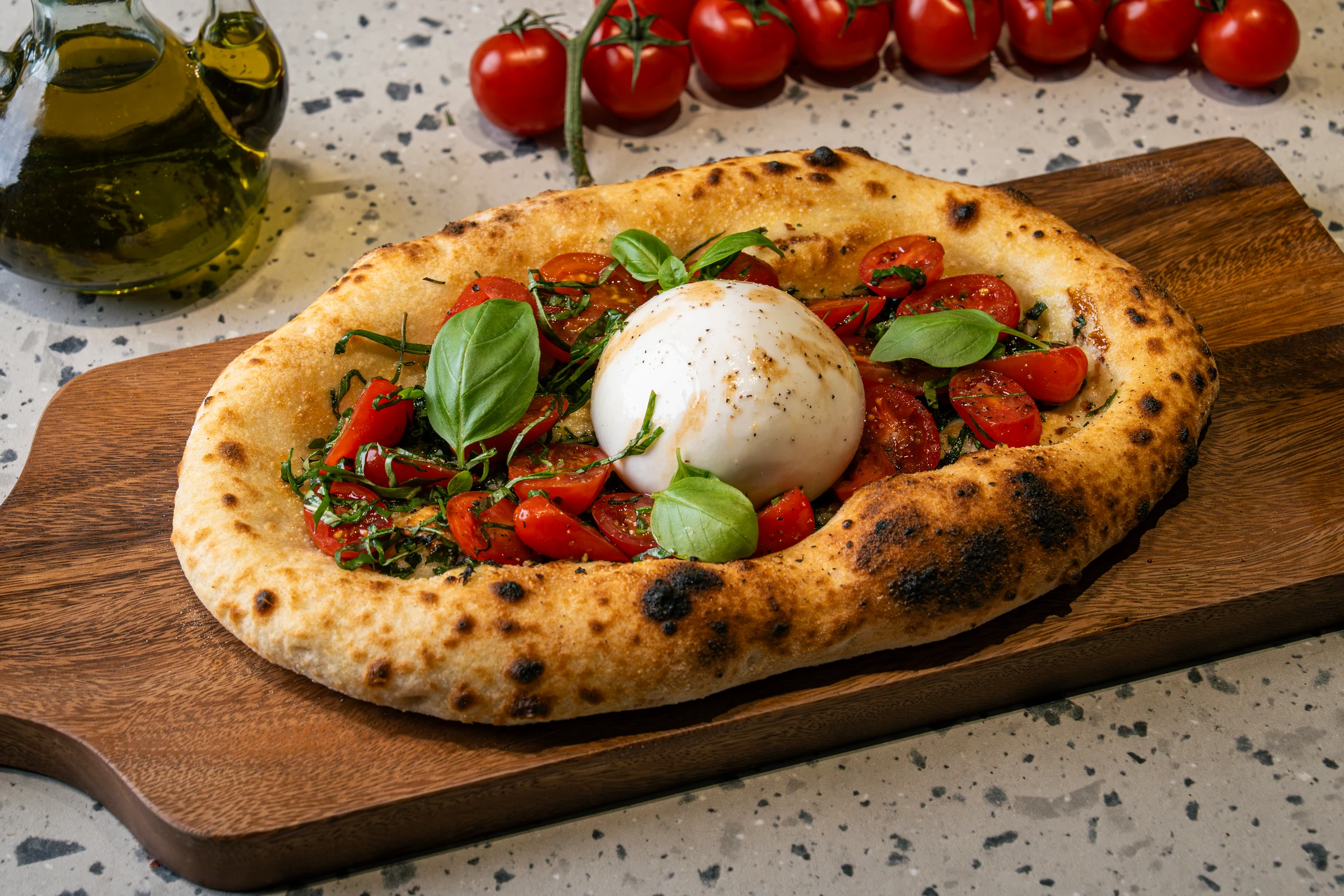 Oval flatbread with a puffed blistered crust, a ball of burrata at the centre, halved cherry tomatoes and shredded basil around it