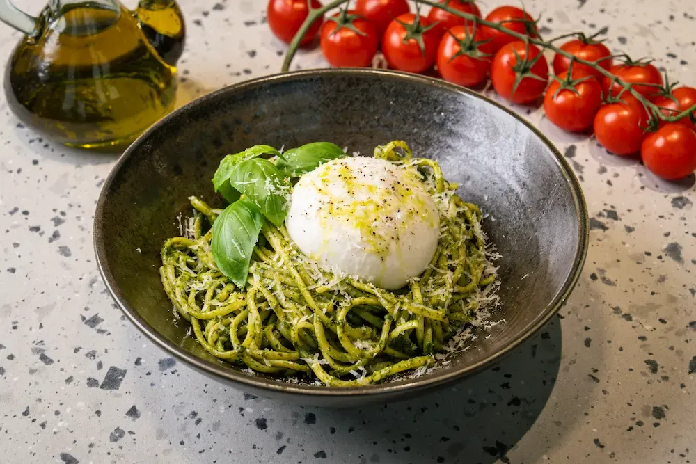 Nest of green pesto linguini in a dark stoneware bowl under a whole burrata ball, with grated parmesan, basil and cracked pepper