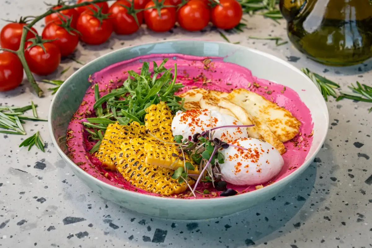 Magenta beetroot purée spread across a shallow bowl under charred corn ribs, chilli-dusted poached eggs, griddled halloumi and rocket