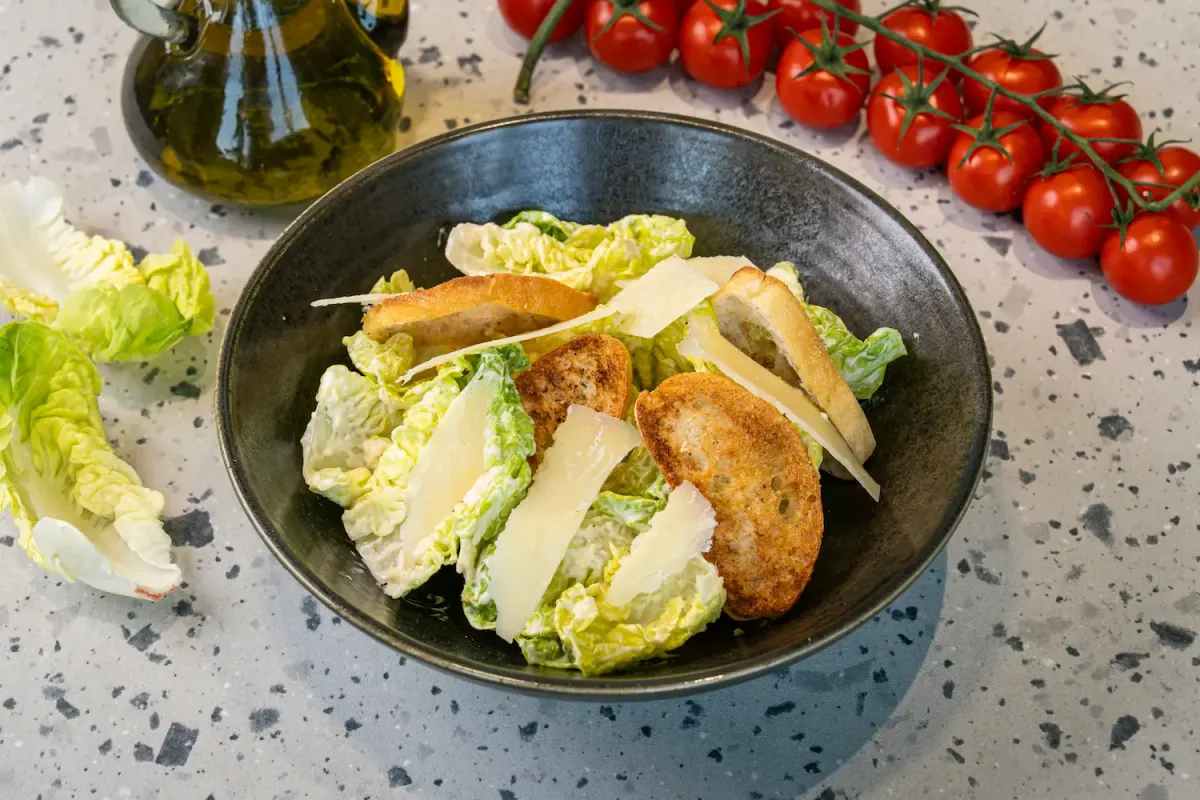 Baby gem leaves in creamy dressing filling a dark charcoal bowl, layered with shaved Parmesan and golden toasted baguette slices