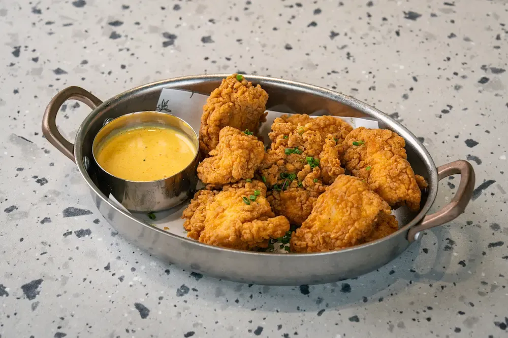 Craggy golden-fried chicken chunks flecked with chives, beside a small steel pot of yellow honey mustard sauce in an oval pan