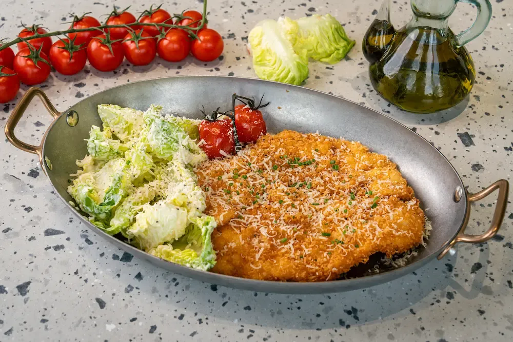 Golden crumbed chicken escalope under grated parmesan and chives in a brass-handled oval pan, beside dressed romaine and blistered tomatoes