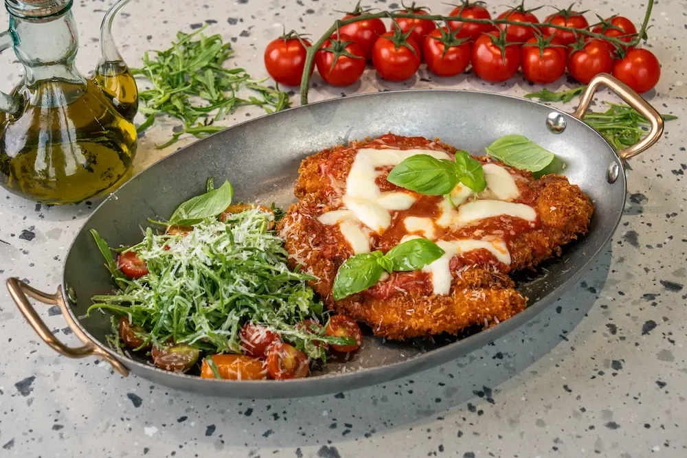 Breaded chicken under tomato sauce and melting mozzarella with basil, in a metal oval pan next to a rocket, tomato and parmesan salad