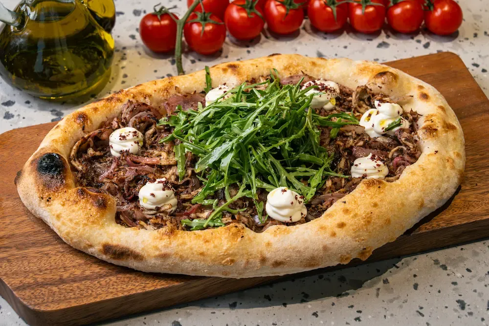 Round flatbread of shredded meat and onions ringed with sumac-dusted labneh dollops, a heap of rocket in the middle, on a wooden board