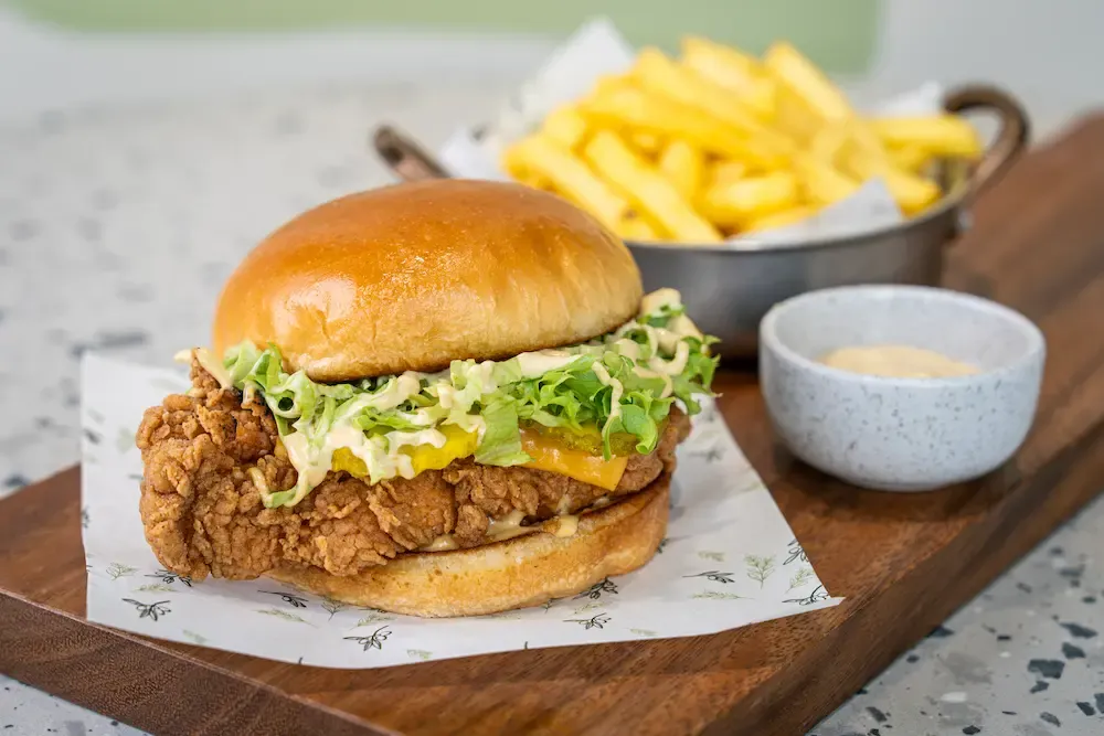 Craggy golden fried chicken fillet with orange cheddar and lettuce in pale sauce, spilling from a brioche bun beside a pot of chips