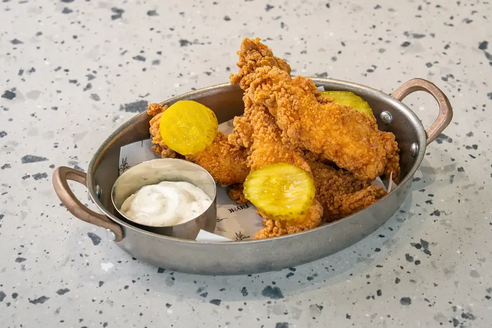Thick craggy fried chicken strips in an oval steel pan with two fat pickle slices and a pot of herb-flecked white ranch