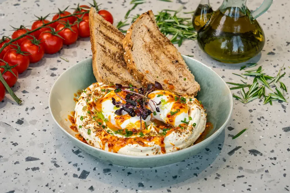 Two poached eggs sunk into whipped labneh with chilli butter, dukkah and chives, griddled seeded toast propped up behind