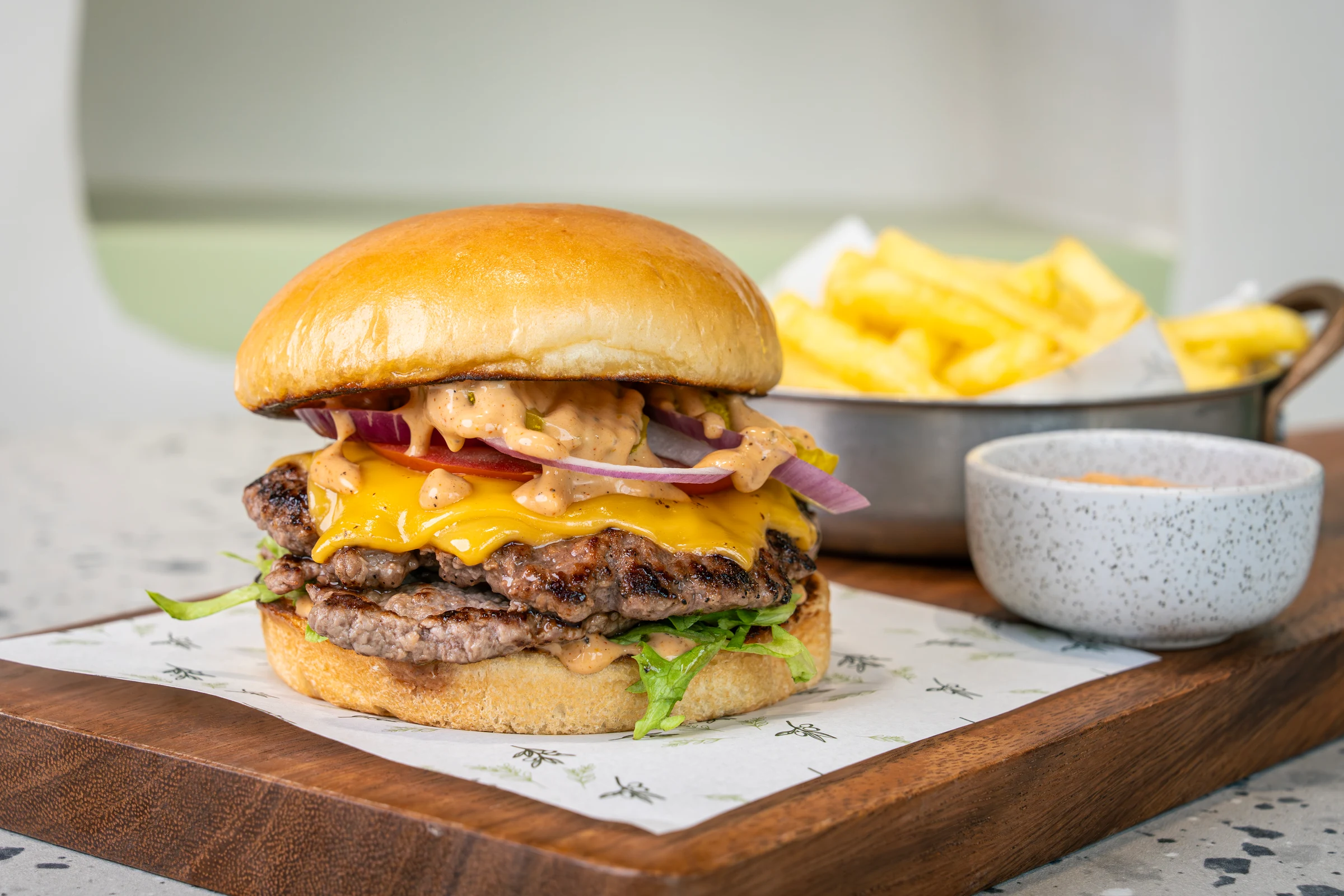 Two griddled patties under melted orange cheddar, tomato, red onion rings and burger sauce in a brioche bun, chips in a metal pot
