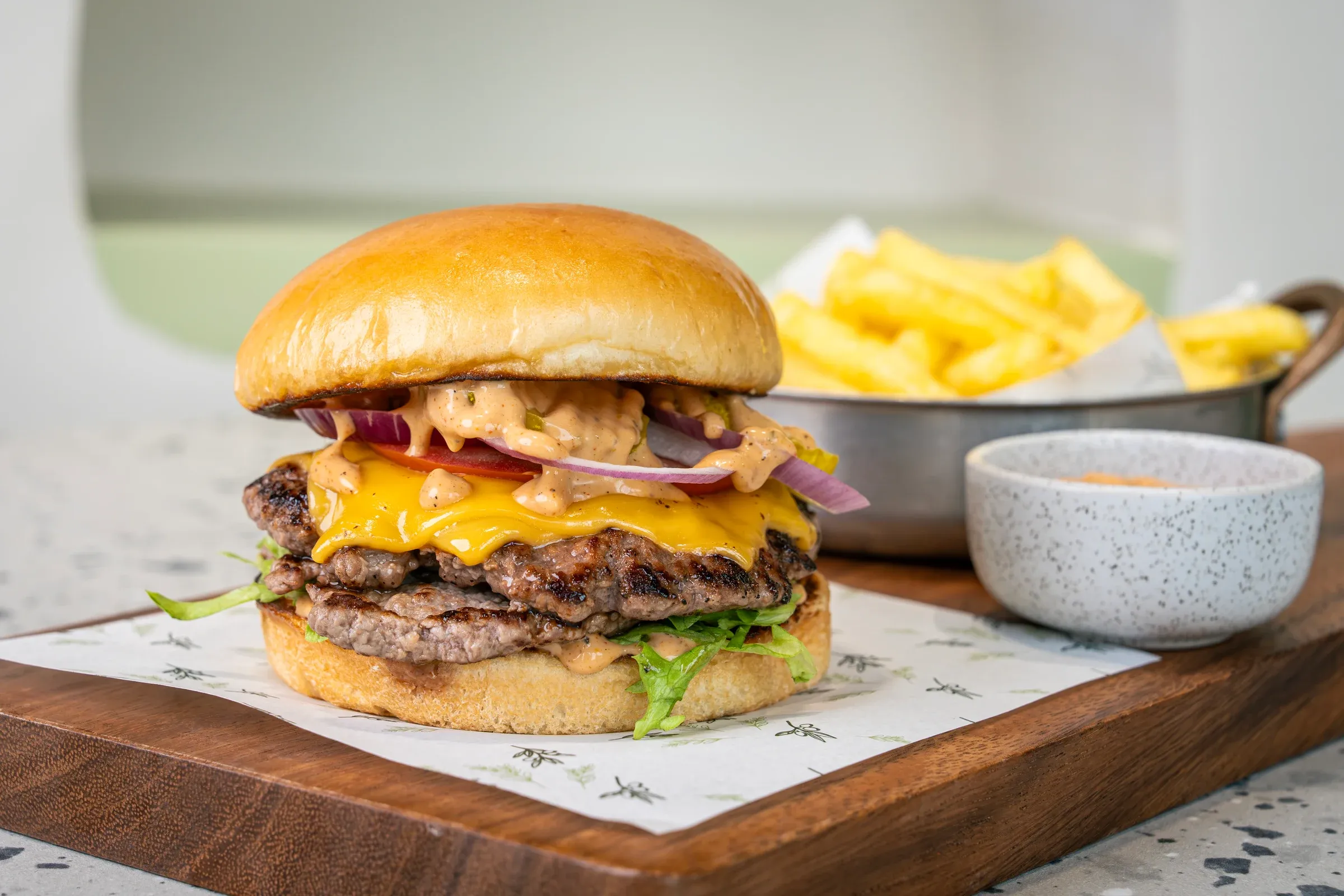 Two griddled patties under melted orange cheddar, tomato, red onion rings and burger sauce in a brioche bun, chips in a metal pot