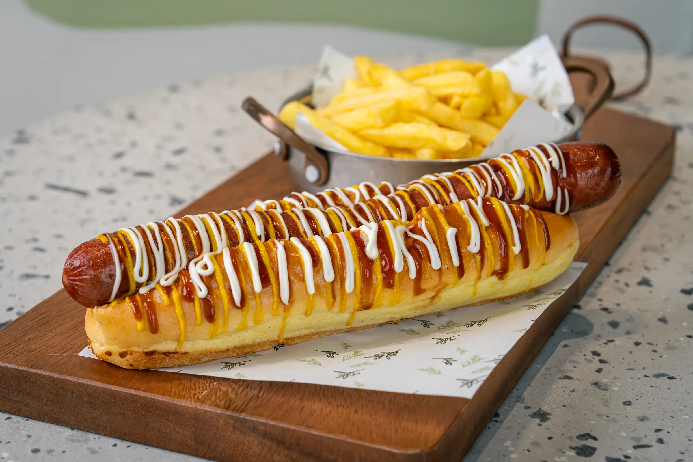 Foot-long sausage overhanging a soft split roll, striped end to end with ketchup, mustard and mayonnaise, chips in a metal pot behind