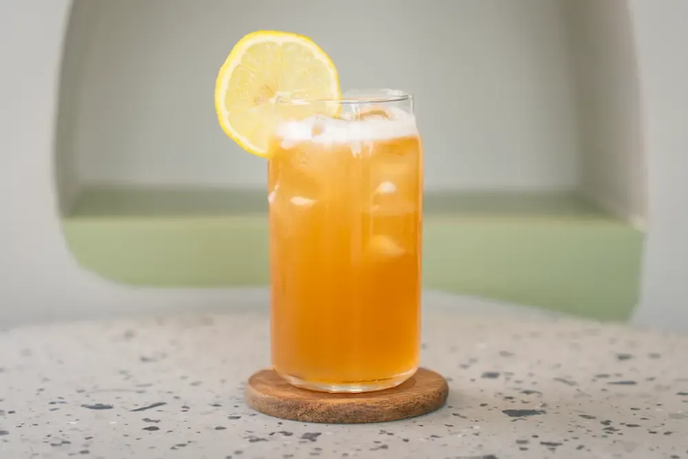 Cloudy amber tea over ice in a can-shaped glass with a foam head and a lemon wheel on the rim, set on a small wooden coaster