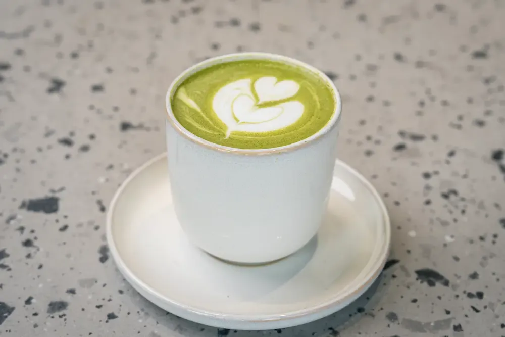 Bright green matcha topped with a white heart in the foam, in a pale glazed tumbler on a matching saucer