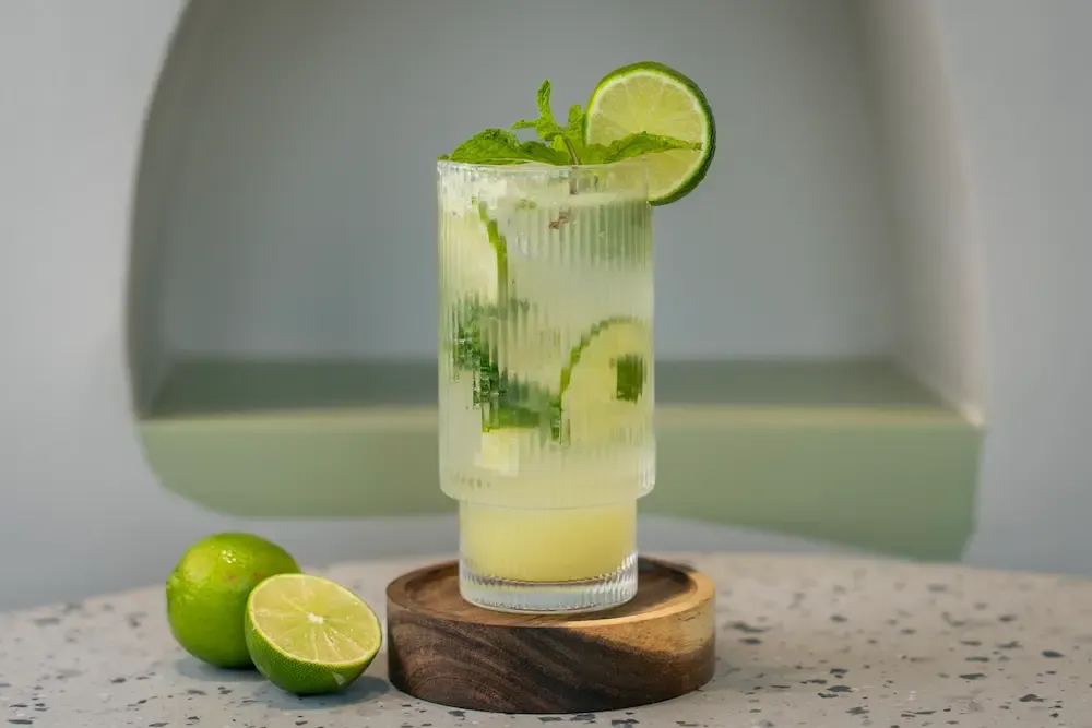 Pale cloudy lime drink in a ribbed glass with mint and a lime wheel on the rim, a whole and halved lime beside the wooden coaster