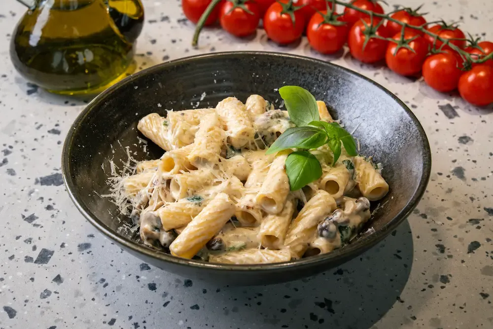 Rigatoni coated in a pale cream sauce with sliced mushrooms and wilted spinach, grated parmesan and a basil sprig, in a dark bowl