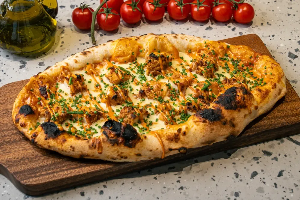 Melted mozzarella under orange sauce zigzags with fried chicken pieces and chopped chives, charred blisters along the puffed rim