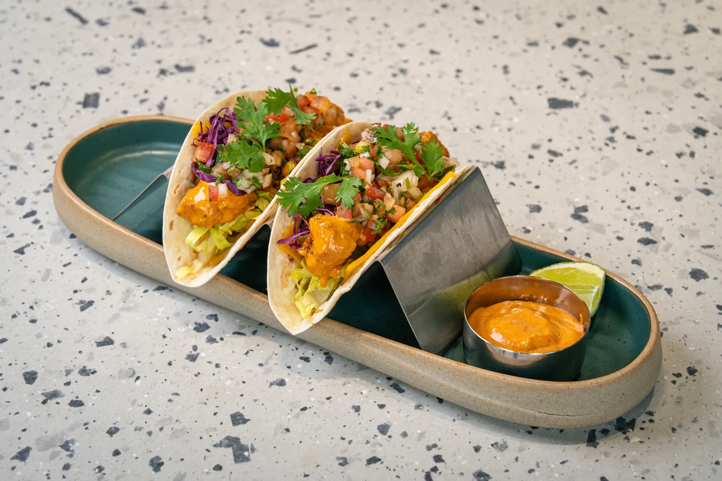Two soft tortillas in a metal stand, filled with orange-glazed chicken, shredded lettuce, purple cabbage and tomato salsa, lime alongside