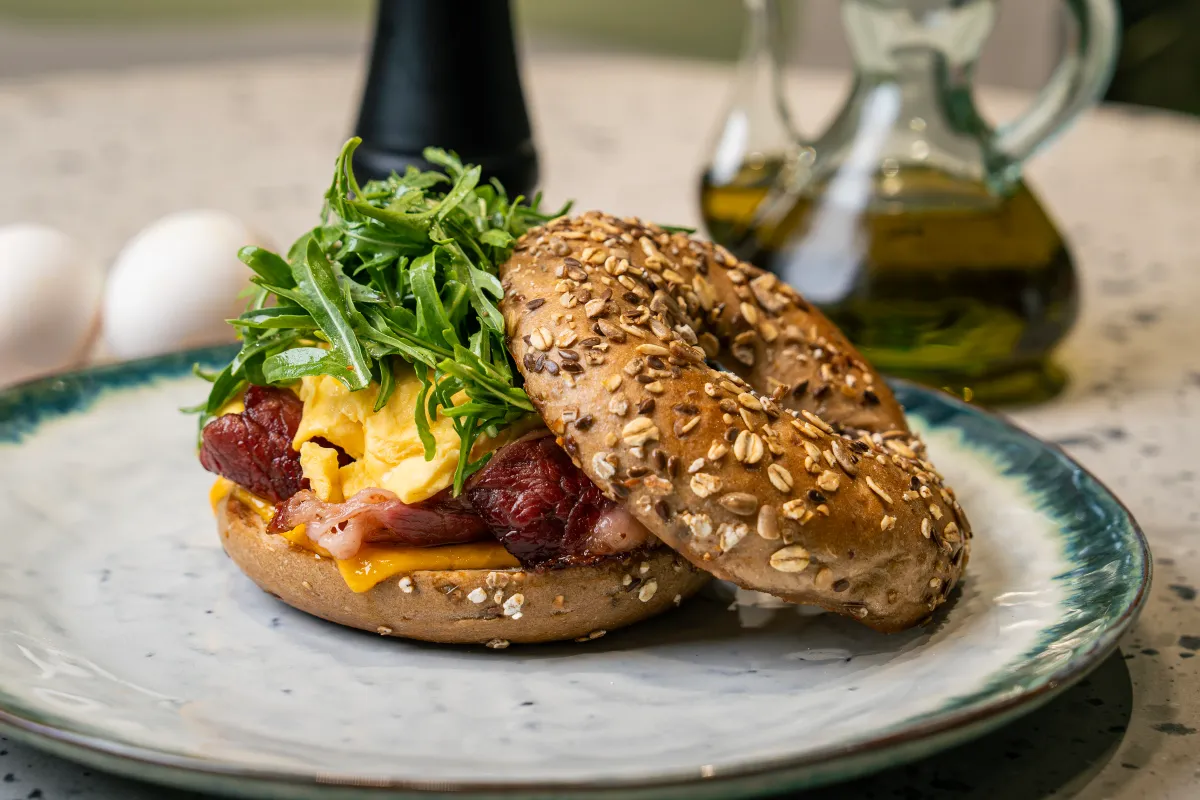 Open seeded bagel with melted cheddar, beef bacon, scrambled egg and rocket, the oat-crusted lid leaning against it