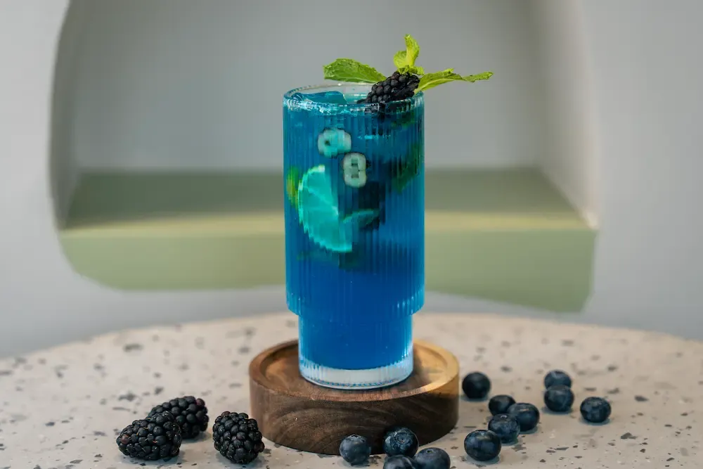 Bright blue drink in a ribbed glass, lime slices and mint suspended inside, blackberry and mint on top, berries scattered around