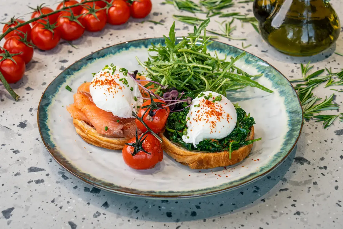 Two croissant halves, one under smoked salmon, one under wilted spinach, each carrying a paprika-dusted poached egg, with rocket alongside