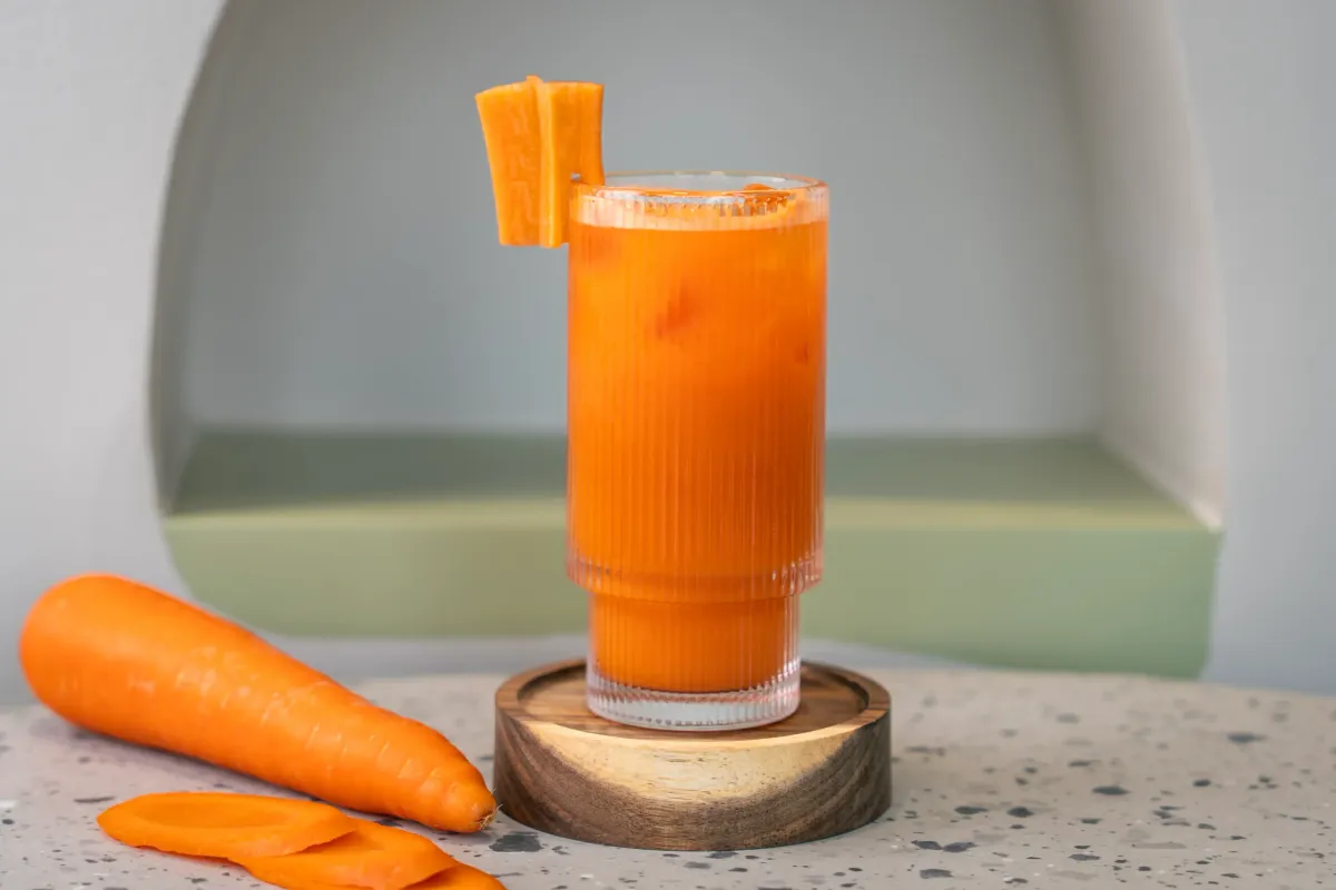 Bright orange juice in a tall ribbed glass, two carrot ribbons perched on the rim, a whole carrot and peeled slices alongside