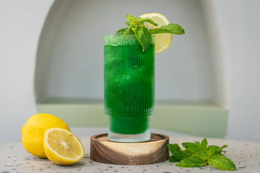 Vivid emerald drink in a tall ribbed glass topped with a mint sprig and lemon wheel, lemons and loose mint leaves alongside