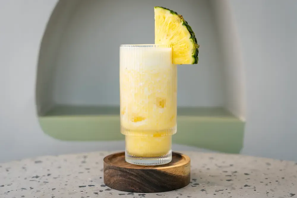 Ribbed glass of pale, pulpy pineapple juice on a round wooden coaster, with a skin-on wedge of pineapple perched on the rim