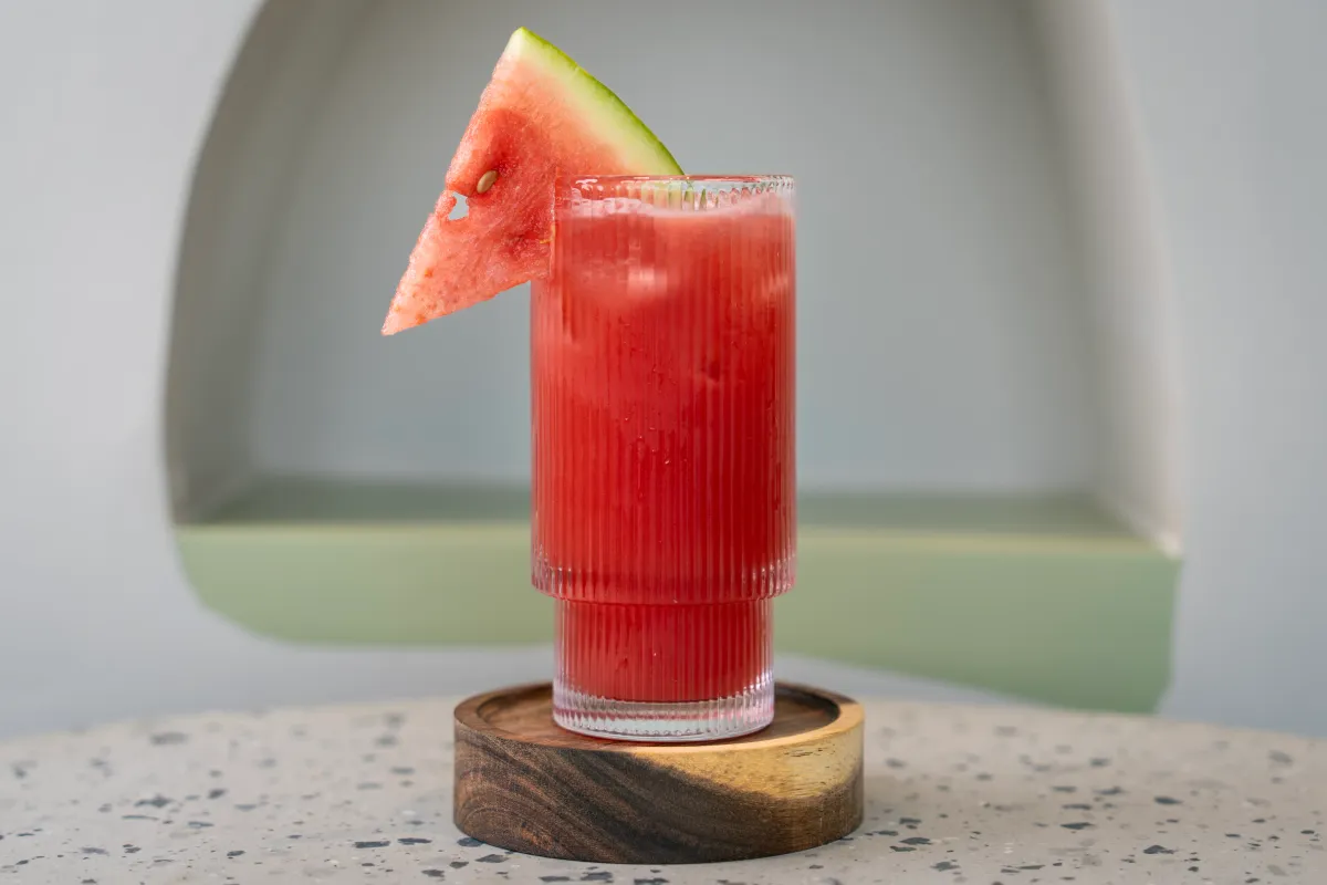 Deep pink watermelon juice in a ribbed glass on a wooden coaster, a rind-edged triangle of melon wedged over the rim