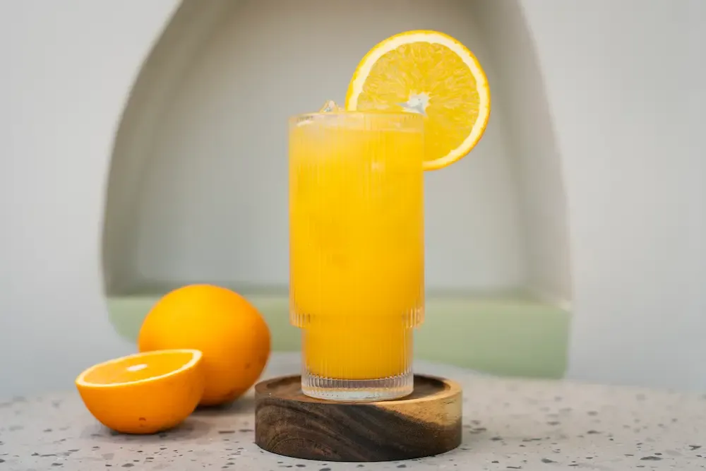 Tall ribbed glass of bright orange juice on a wooden coaster, a wheel of orange notched over the rim and a halved orange beside it