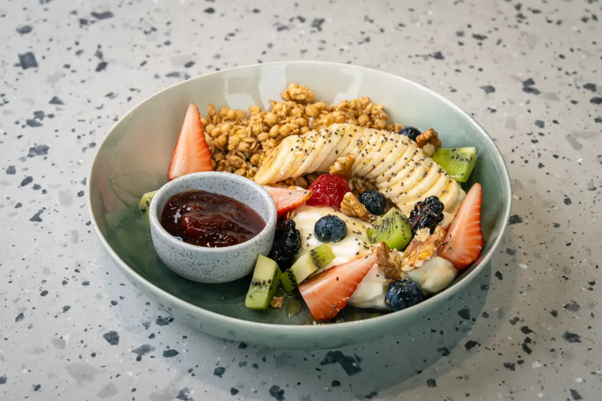 Granola clusters, honeyed banana fanned with chia seeds, berries, kiwi and walnut halves over yoghurt, with a small pot of red jam