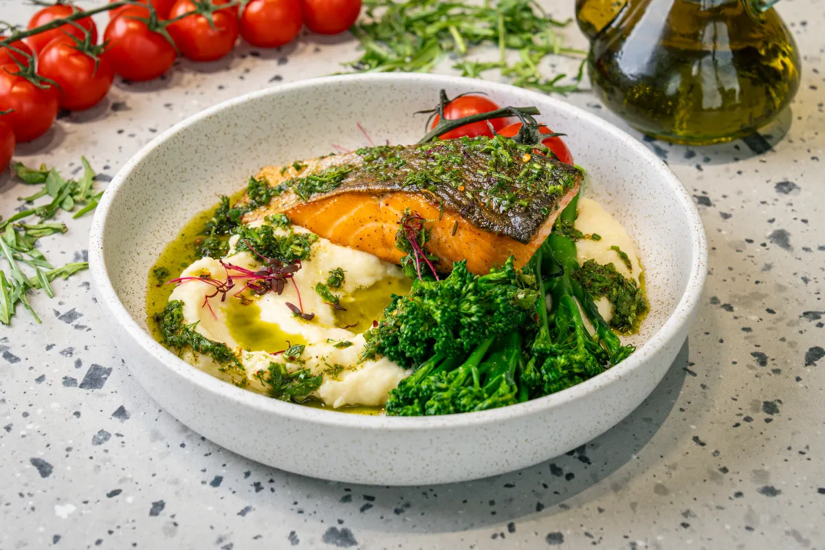 Crisp-skinned salmon crusted with herbs on whipped mash, chimichurri oil pooling around deep green broccolini and red micro leaves