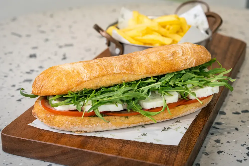 Long crusty baguette split open over thick white halloumi slices, tomato, green pesto and a mound of rocket spilling out, chips behind