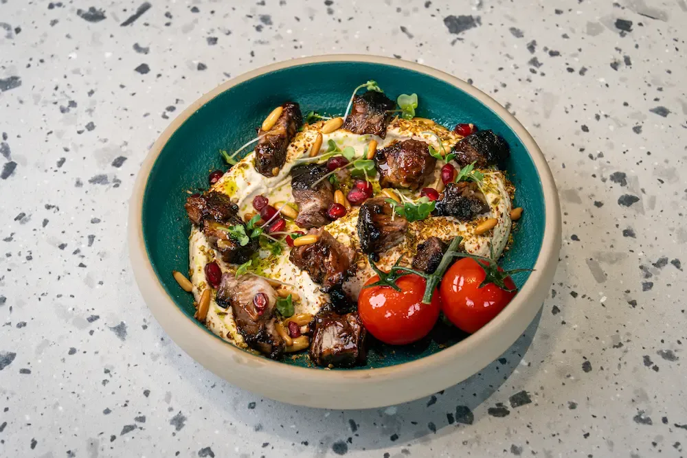 Dark caramelised beef pieces over smooth hummus in a teal bowl, with pine nuts, pomegranate seeds and two tomatoes on the vine
