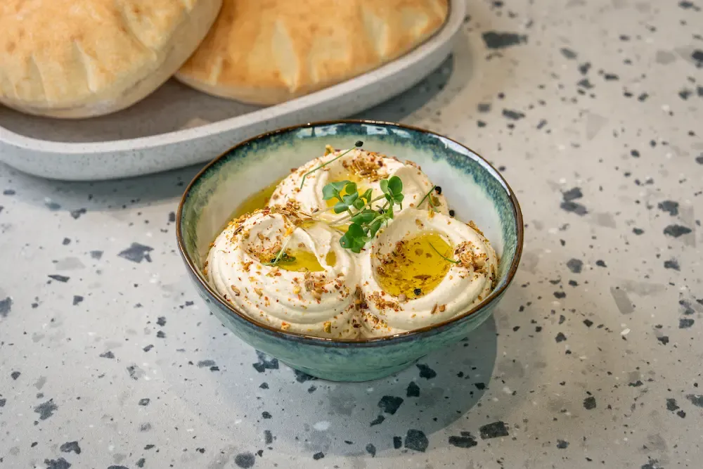 Whipped pale hummus swirled around a pool of olive oil, dusted with za'atar and crowned with micro greens; warm pitta behind