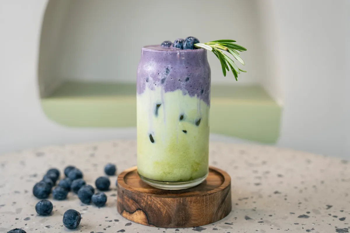 Tall glass layered pale green under deep purple foam, whole blueberries and a rosemary sprig on the rim, loose berries below
