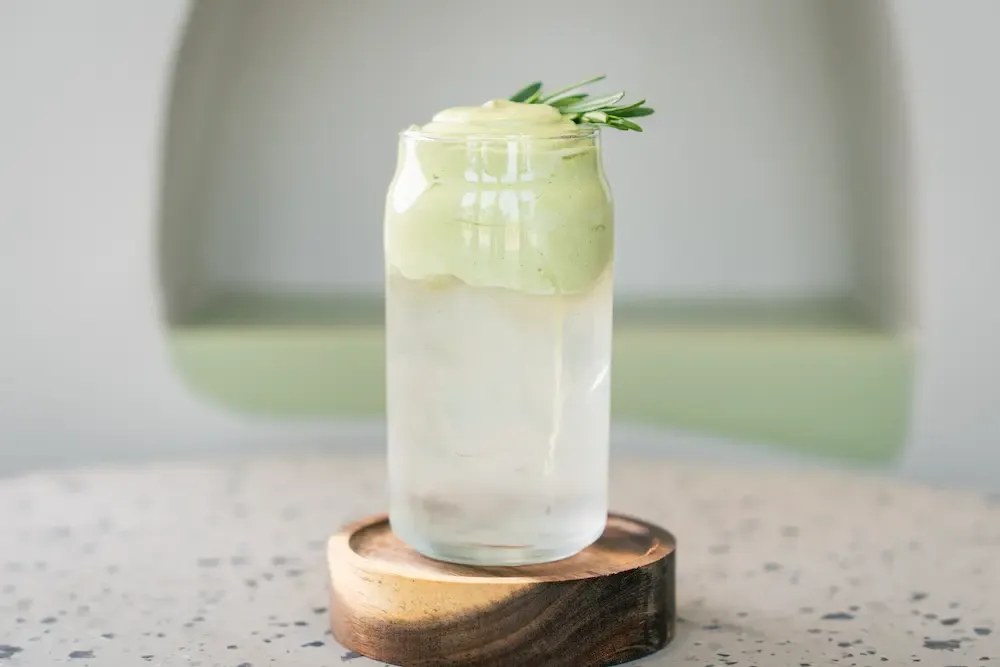 Clear glass of ice capped with thick pale green foam that spills over the rim, a rosemary sprig tucked alongside