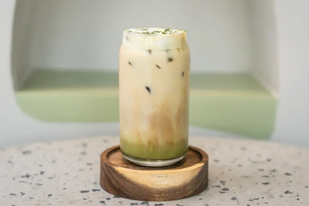 Tall can-shaped glass on a round wooden coaster, a green pistachio layer settled under milky coffee and a foam cap dusted green